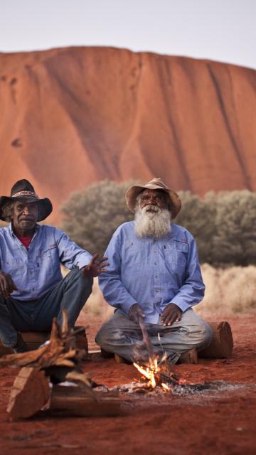 Two Anangu men sit by a fire